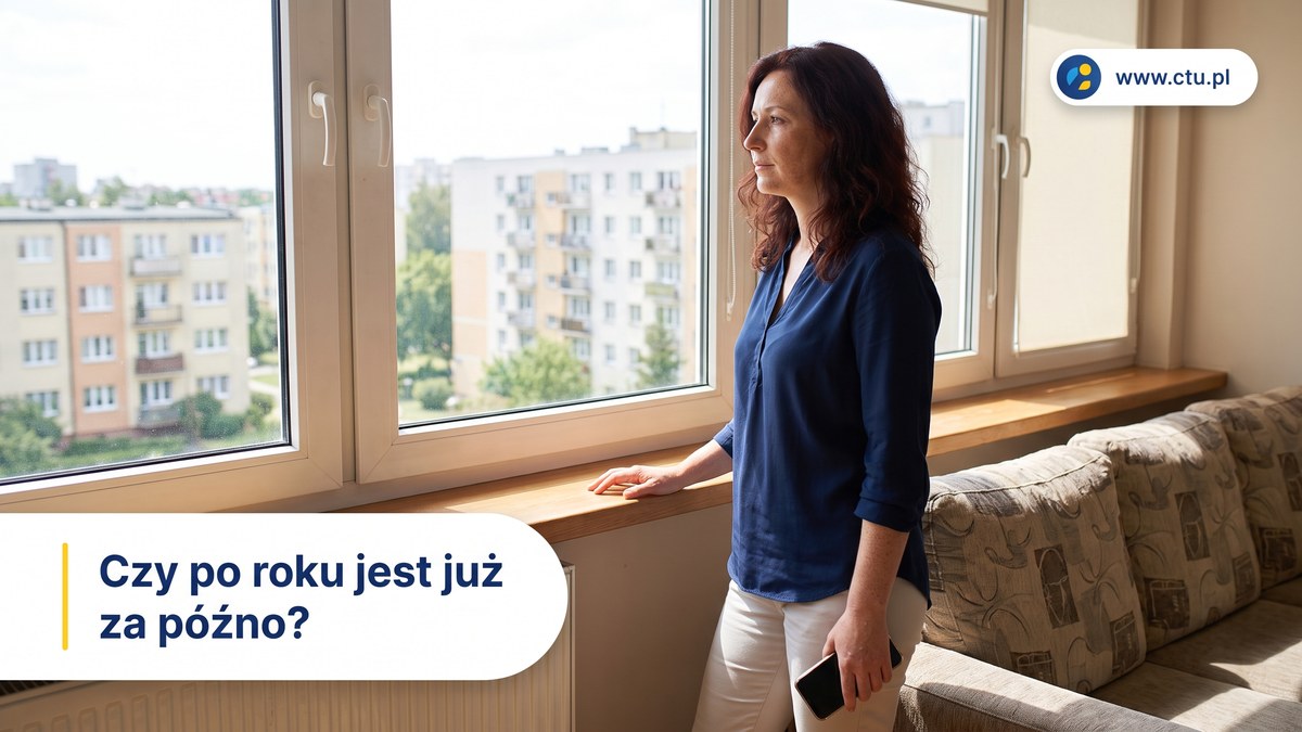Kobieta stoi przy oknie mieszkania z telefonem w opuszczonej dłoni i patrzy na osiedlową zabudowę.
